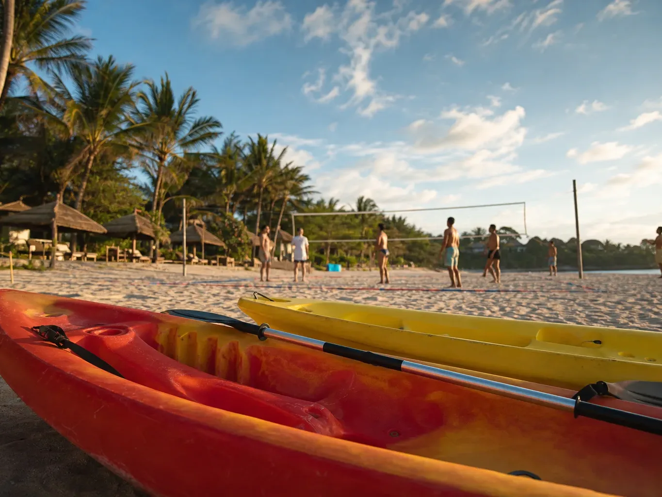 Two kayaks rest on pale sandy beach in the foreground whilst a group of people play volleyball on a net court behind them, surrounded by palm trees and resort huts