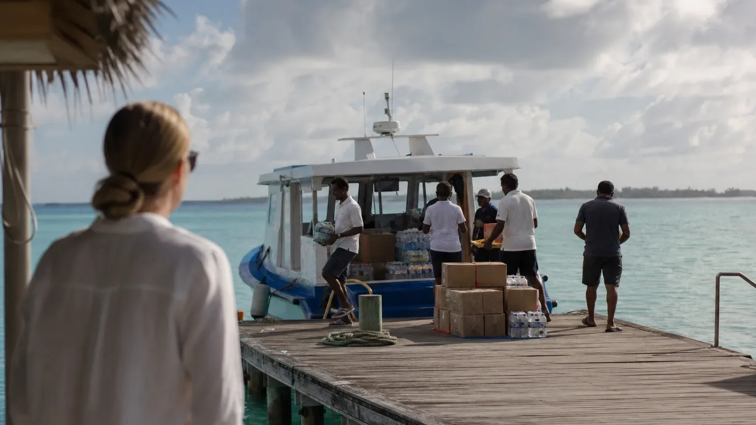 A person watches from a wooden jetty over turquoise water as a supply boat is unloaded, with cardboard boxes and cases of bottled water stacked on the dock