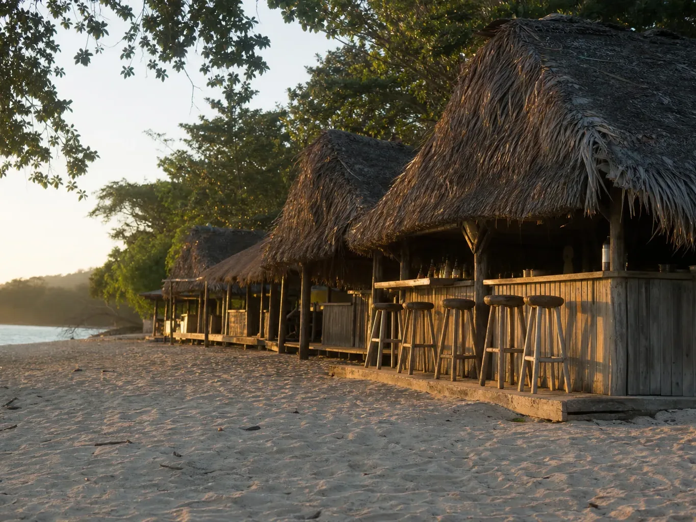 A row of thatched open-air wooden beach bars lines a sandy shore at golden hour, bar stools empty along the rustic timber counters beneath palm-frond roofs