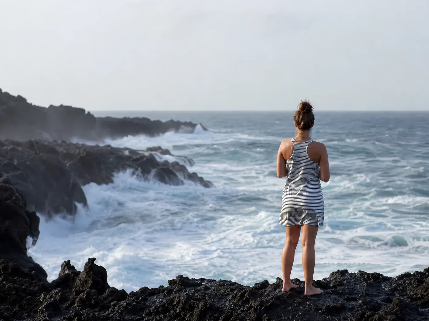 A person in a striped sleeveless dress stands barefoot on dark volcanic rocks, looking out over a churning ocean with crashing waves