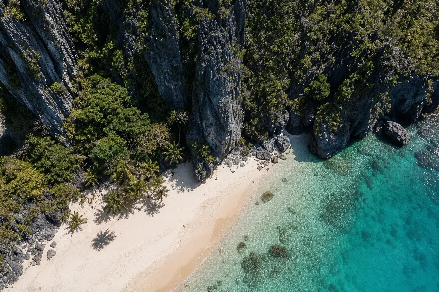 An aerial view of a deserted tropical beach of pale sand edged by turquoise shallow water and backed by tall dark limestone cliffs draped in dense green vegetation