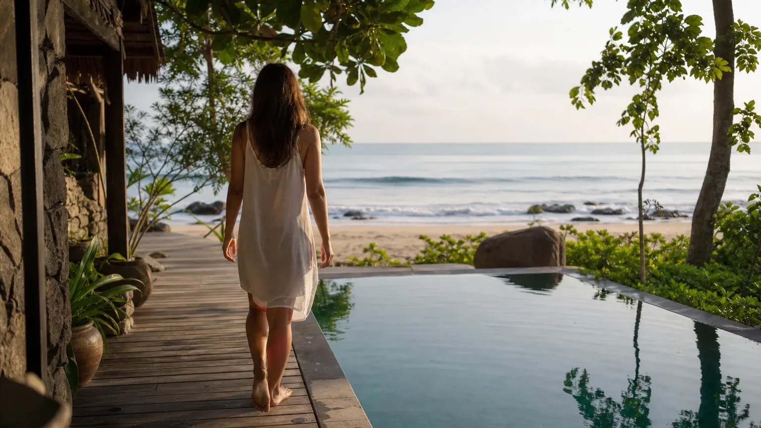 A person in a white dress walks barefoot along a timber deck beside a plunge pool, with an open sandy beach and calm sea ahead