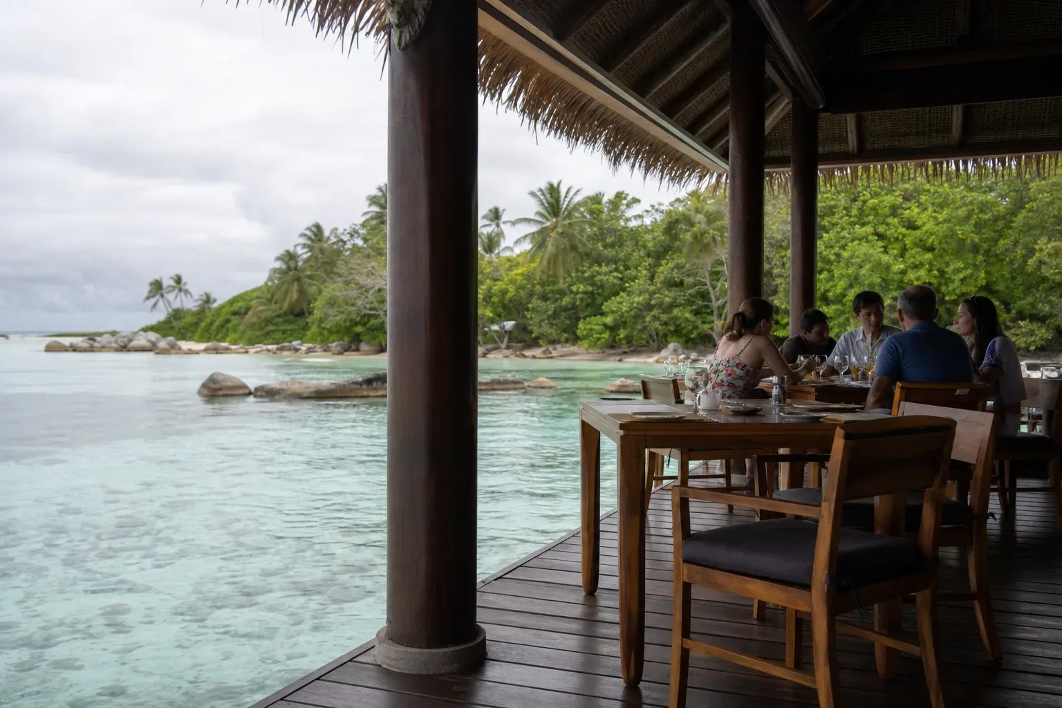 A group of guests dine at wooden tables on an open-air overwater terrace with a thatched roof, overlooking a calm turquoise lagoon fringed with lush tropical vegetation