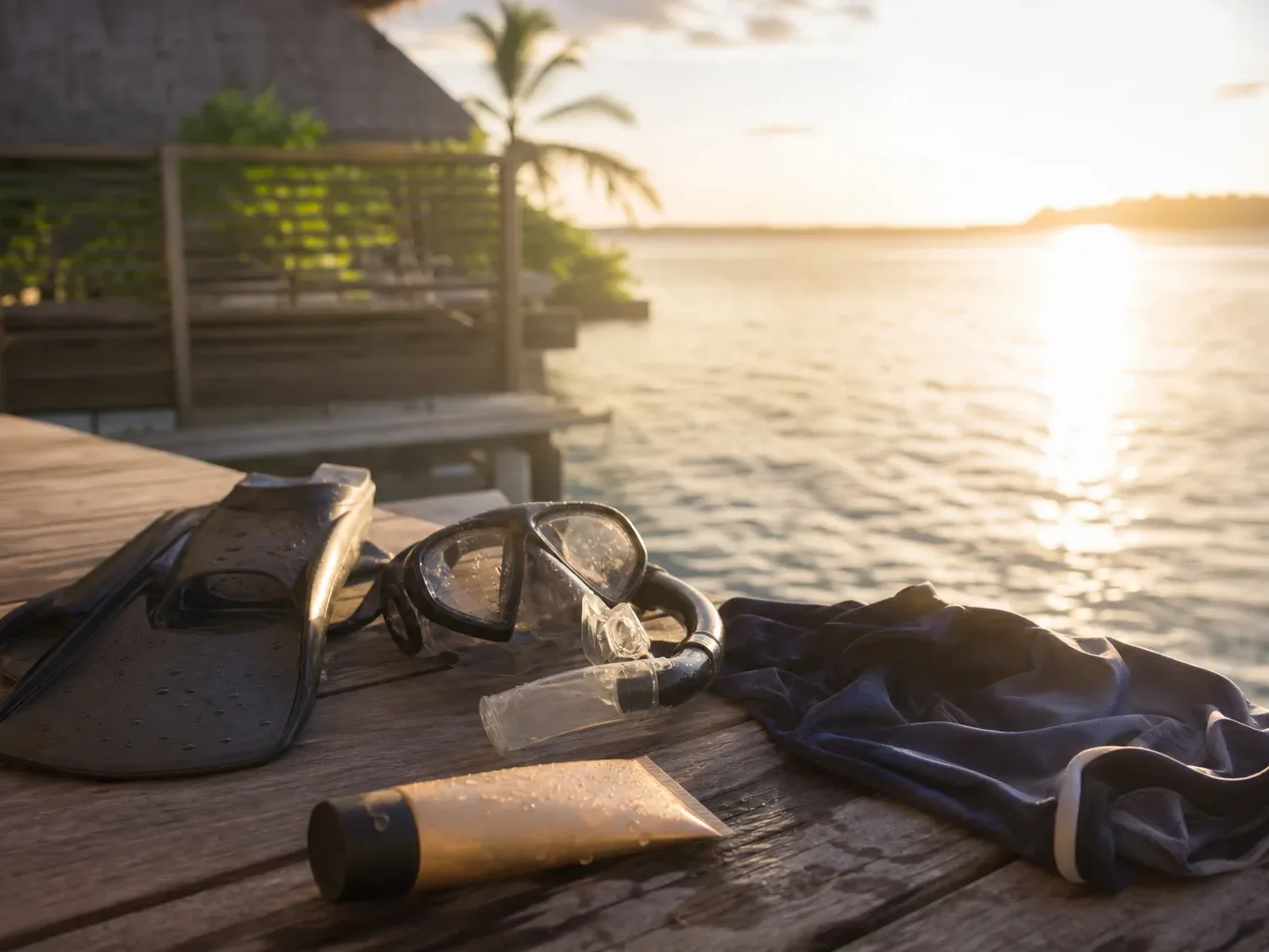 Snorkelling fins, a dive mask with snorkel, a tube of lotion, and a dark rash vest laid on a wet wooden deck beside a calm lagoon at golden hour, with an overwater villa and palm trees behind
