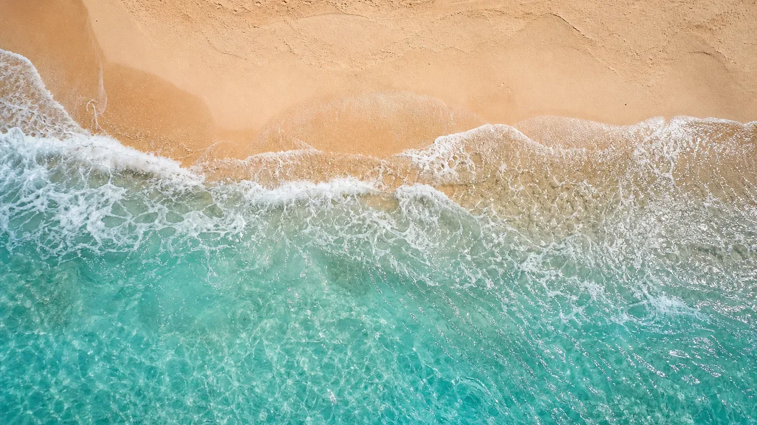 Turquoise shallow water breaks in white foam along a pale sandy shore, photographed from directly overhead