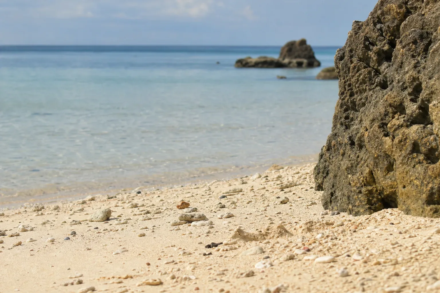 A sandy beach beside a pitted limestone rock formation, with clear shallow turquoise water and a rocky outcrop in the distance under a partly cloudy sky