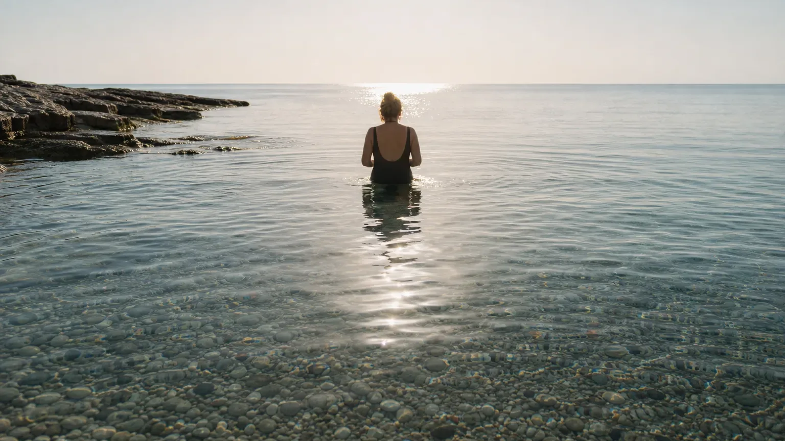 A person in a black swimsuit stands waist-deep in clear, calm sea water beside a rocky shoreline, facing towards the sunlit horizon