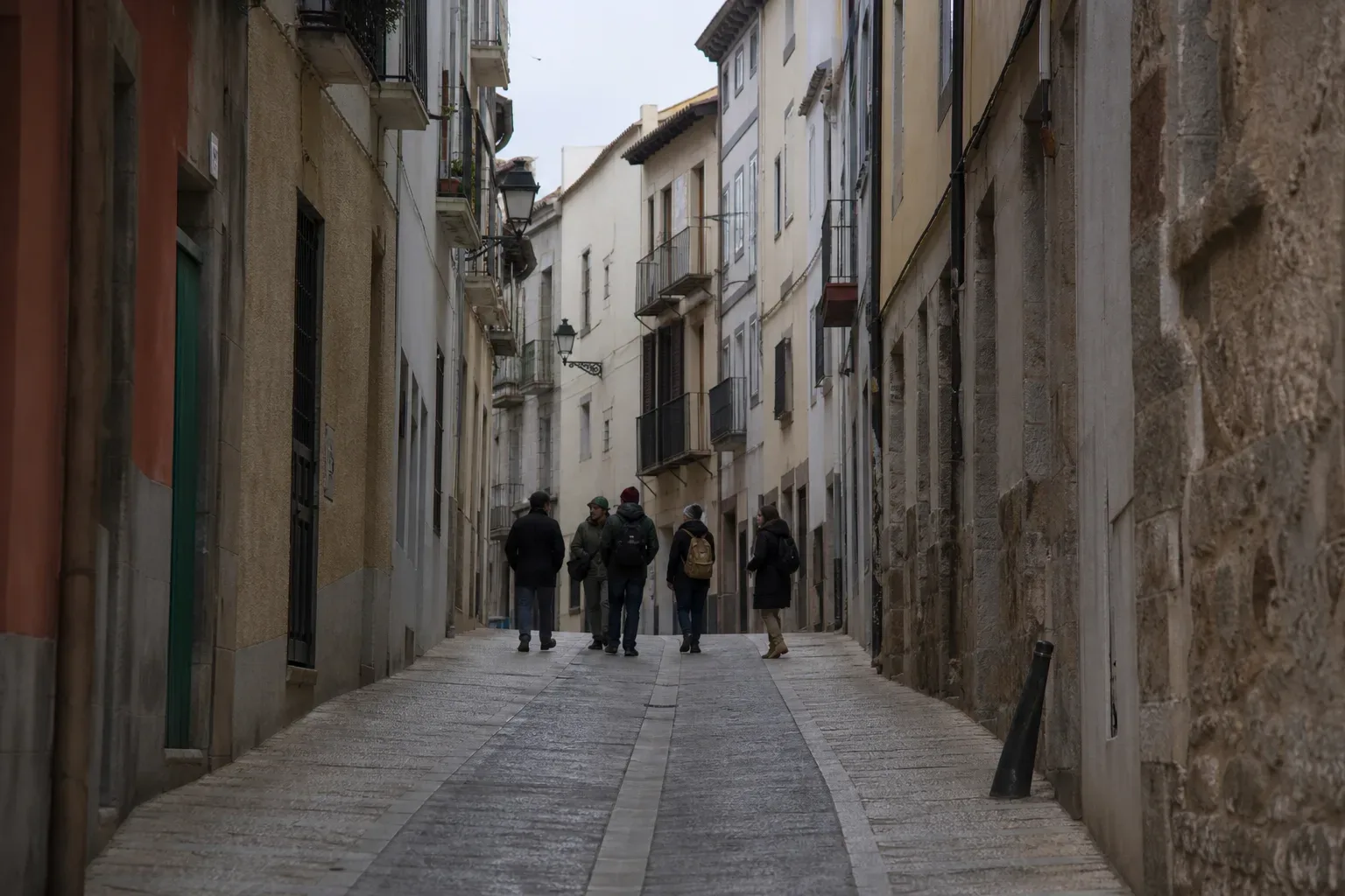 A small group of people walking away along a narrow cobbled street lined with multi-storey buildings and wall-mounted lanterns under flat overcast light