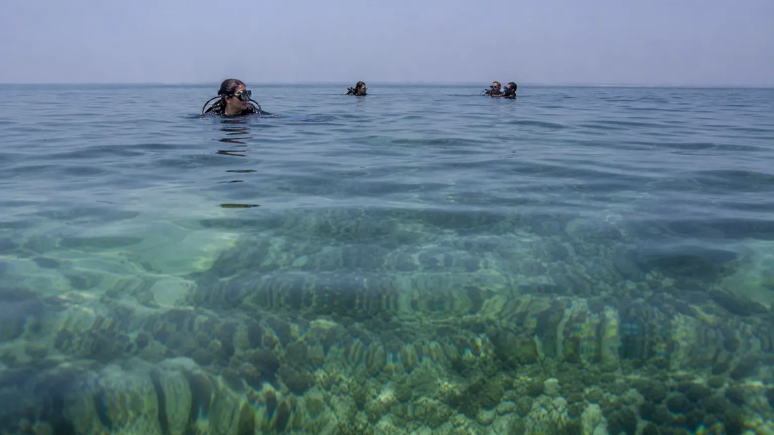 Four scuba divers float at the surface of clear tropical water above a shallow coral reef visible through the calm sea