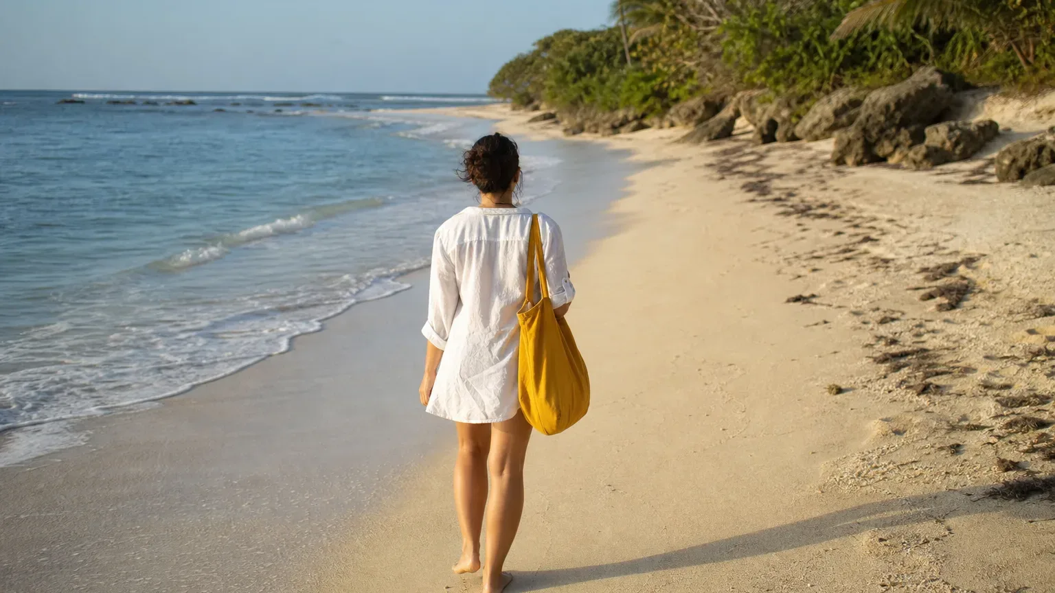 A person in a white shirt and carrying a yellow bag walks barefoot along a pale sandy beach beside calm turquoise water, with lush tropical vegetation lining the shore ahead
