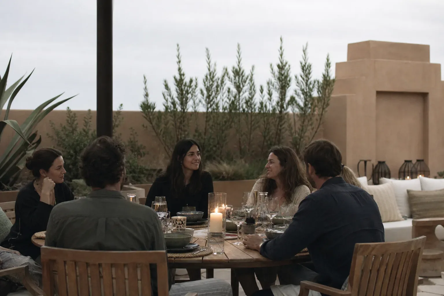 A small group of people seated around a candlelit wooden table on an open outdoor terrace, sharing an evening meal amid desert-style architecture and tall slender trees