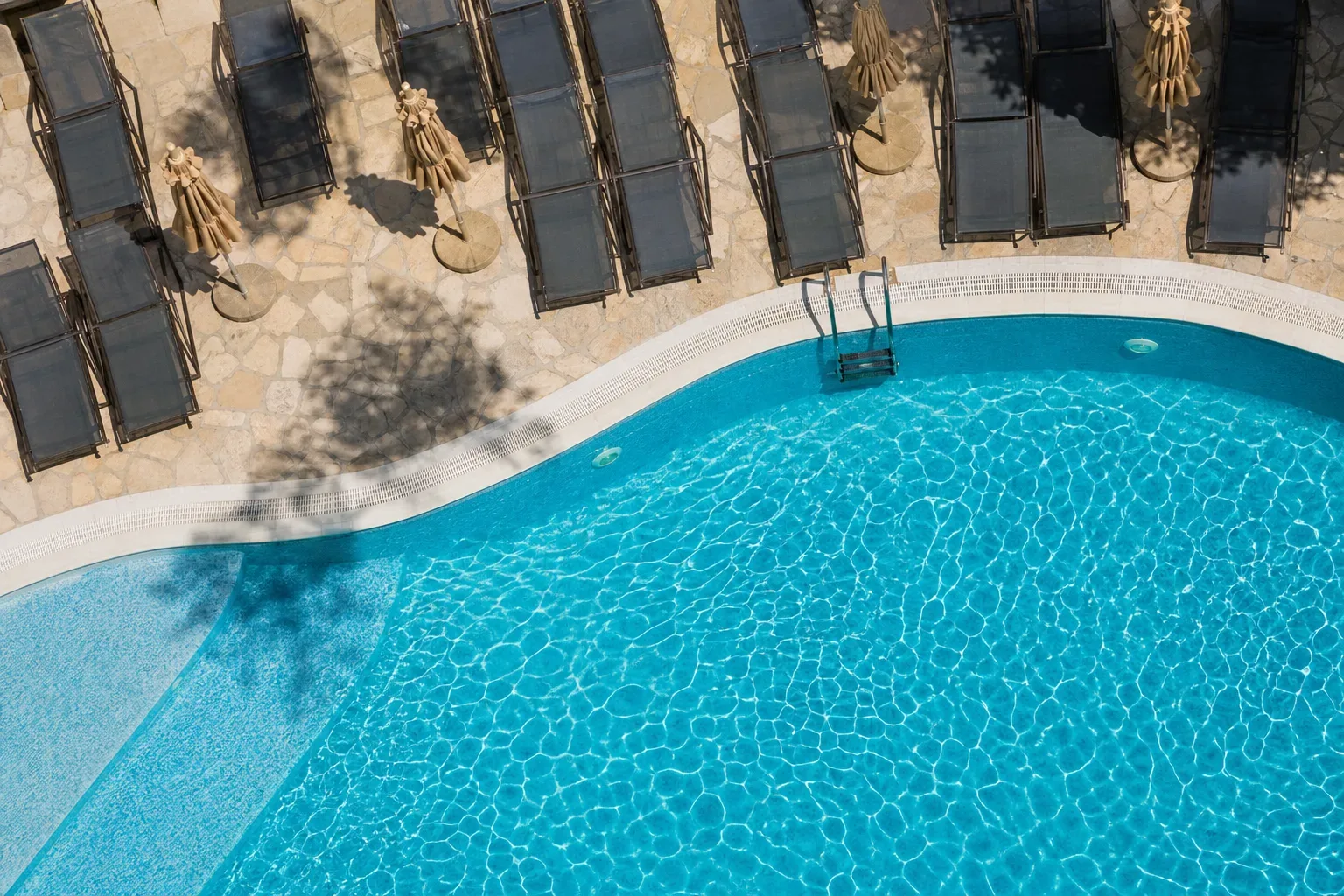 An aerial view of a curved resort swimming pool with vivid turquoise water, surrounded by a stone terrace lined with empty sun loungers and closed parasols