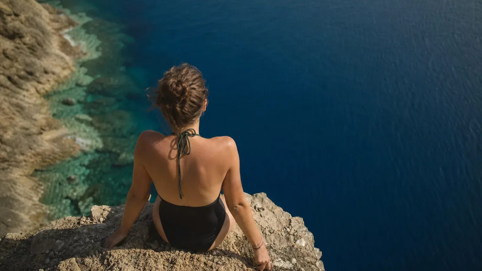 A person in a black swimsuit sits on a rocky cliff edge looking out over deep blue open water far below
