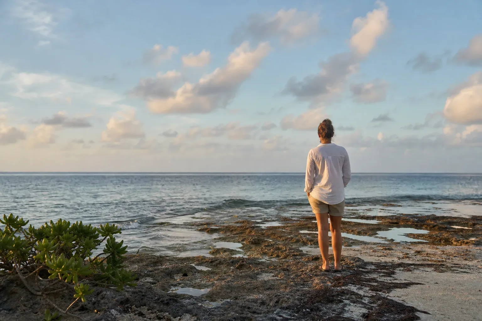 A person stands barefoot on a rocky coral shoreline, looking out across calm open ocean beneath a partly cloudy sky in soft warm light