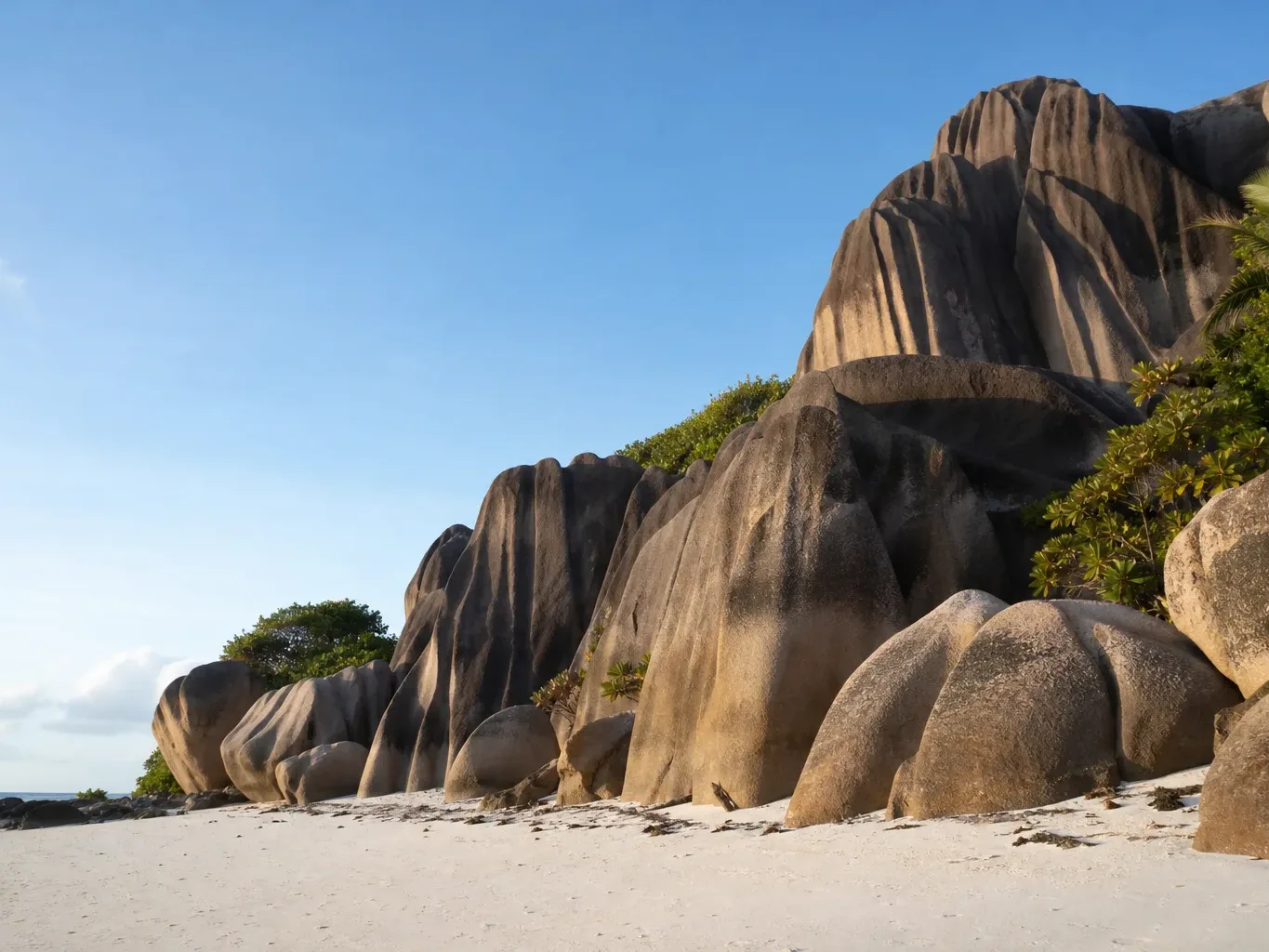 Huge smooth granite boulders, darkened with weathering, rise steeply behind a wide white-sand beach, with green tropical shrubs tucked between the rocks under a clear blue sky