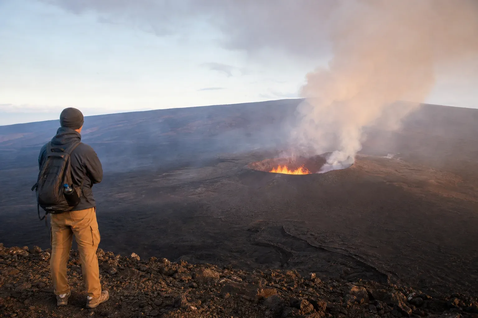A person wearing a backpack stands on the edge of a vast volcanic crater, watching lava burn and smoke billow from an active vent below