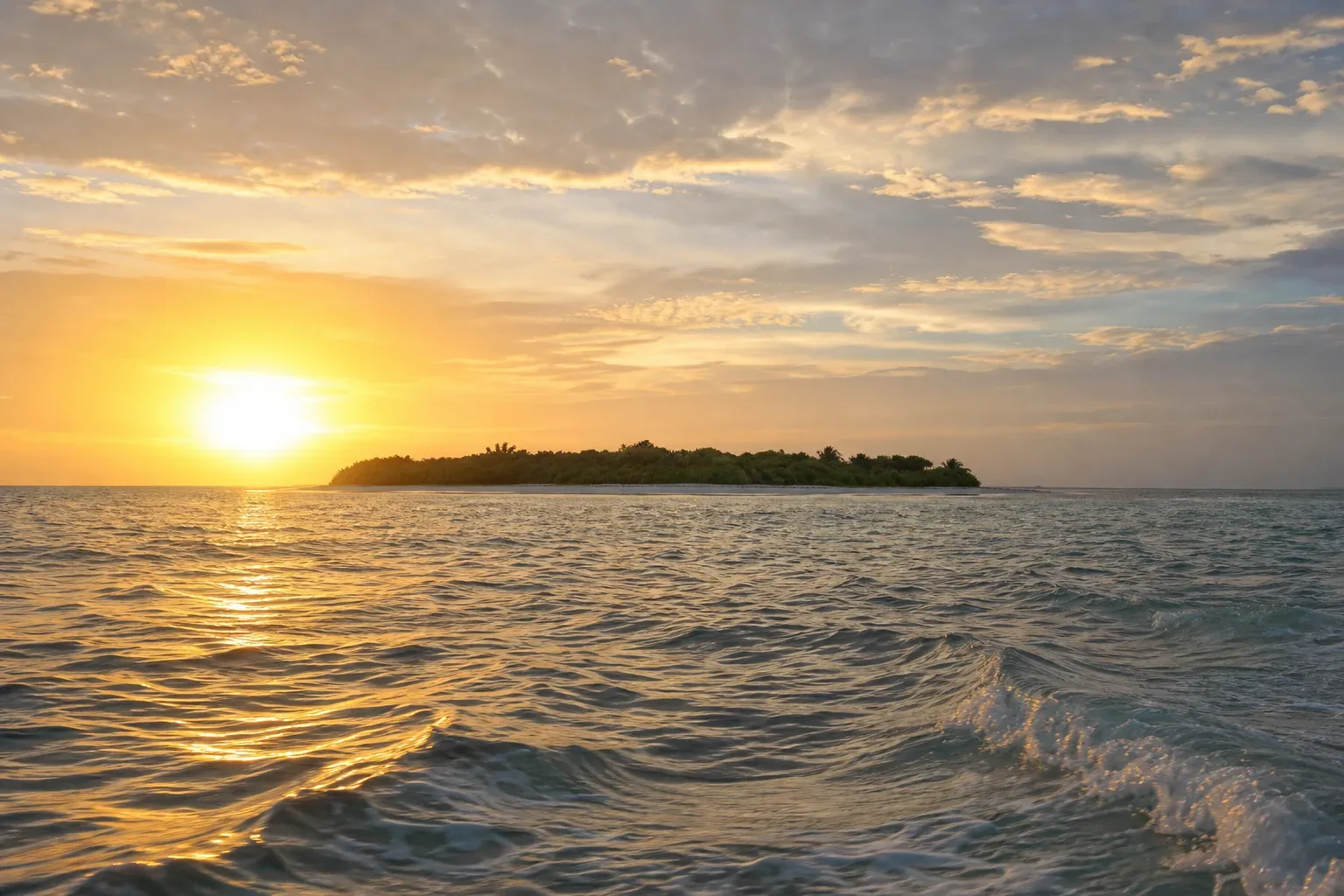 A small tree-covered island silhouetted against a golden sunset over gently rippling open ocean, viewed from water level