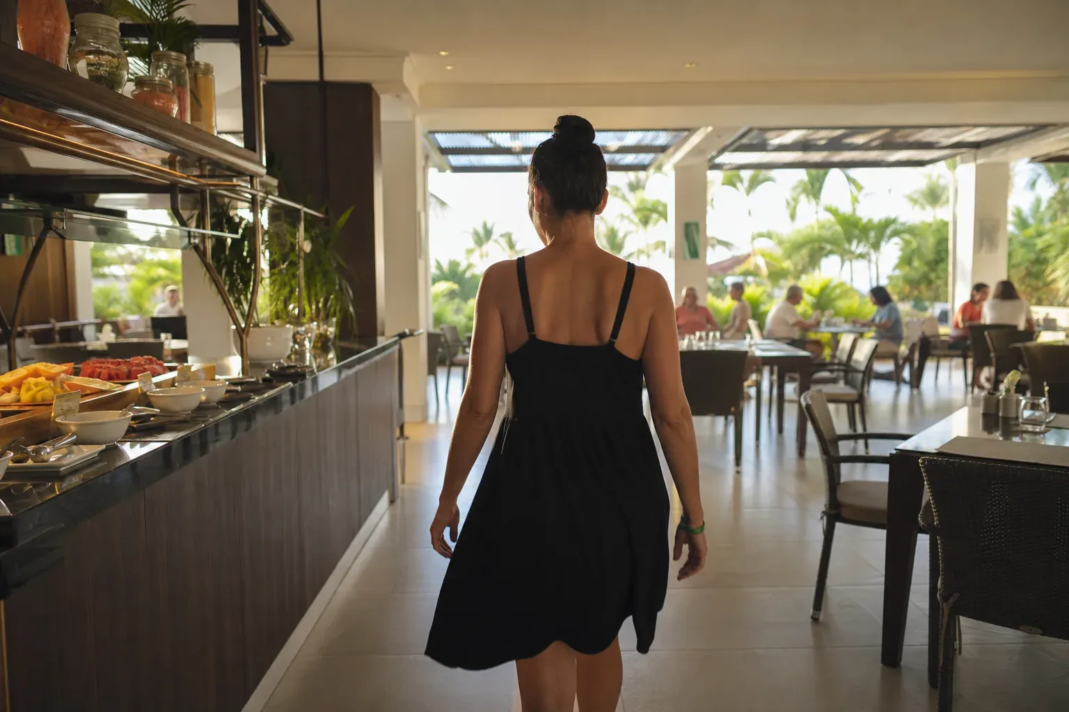 A person in a black sundress walks away from the camera through an open-sided resort buffet dining area, with tropical foliage visible on the terrace beyond