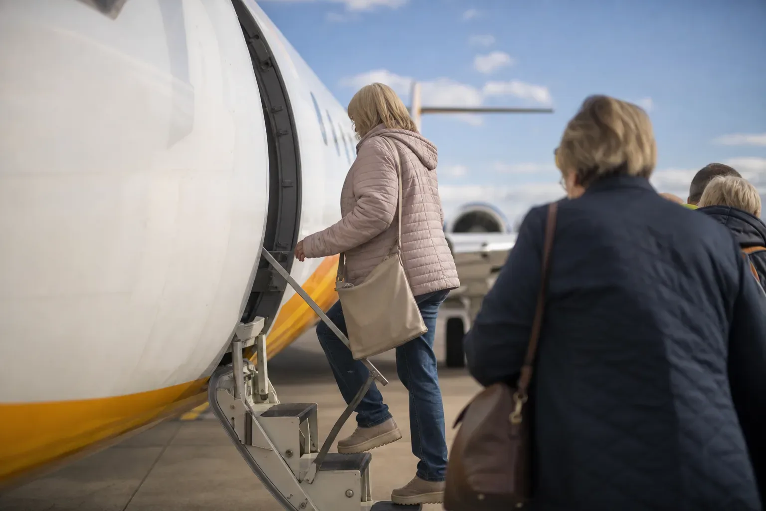 Passengers board a small jet aircraft via external steps on an open apron under a bright blue sky