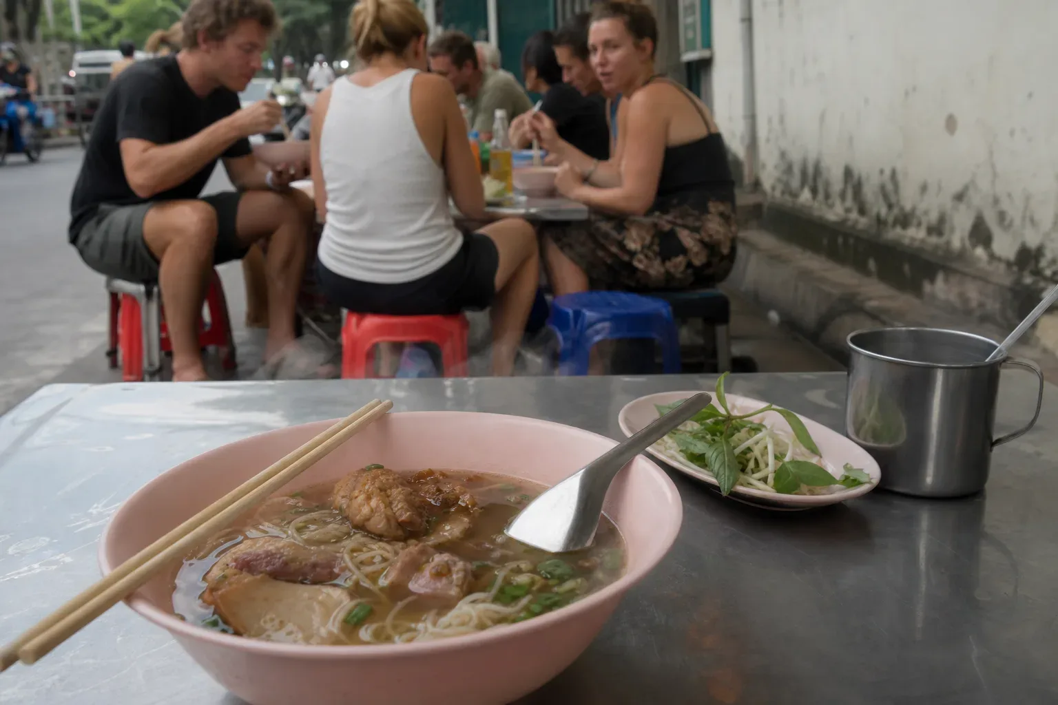 A large bowl of noodle soup with chopsticks and a spoon sits on a metal pavement table, with a side plate of fresh herbs nearby and a group of people eating at low stools in the background