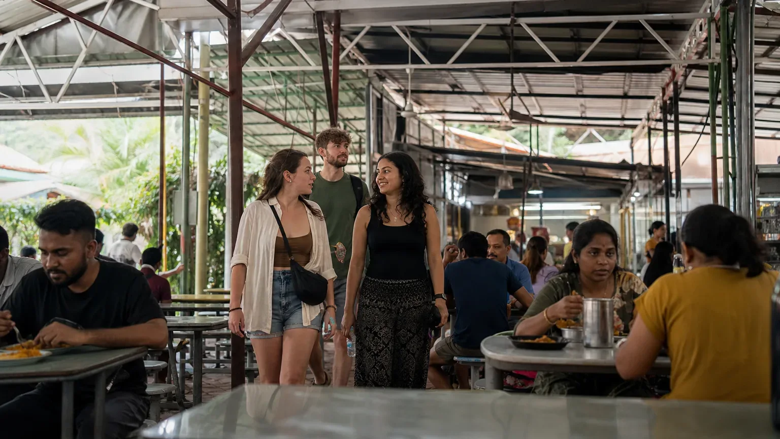 Three people walk through a busy covered outdoor food court with metal roof trusses, while others sit eating at nearby tables