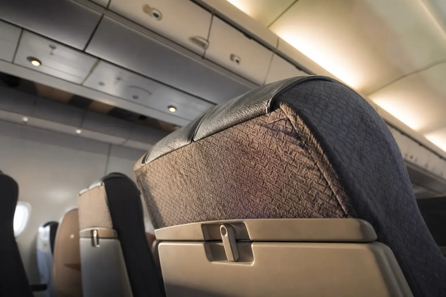 Close-up of an empty economy aircraft seat headrest and folded tray table, with overhead luggage bins and cabin ceiling visible behind