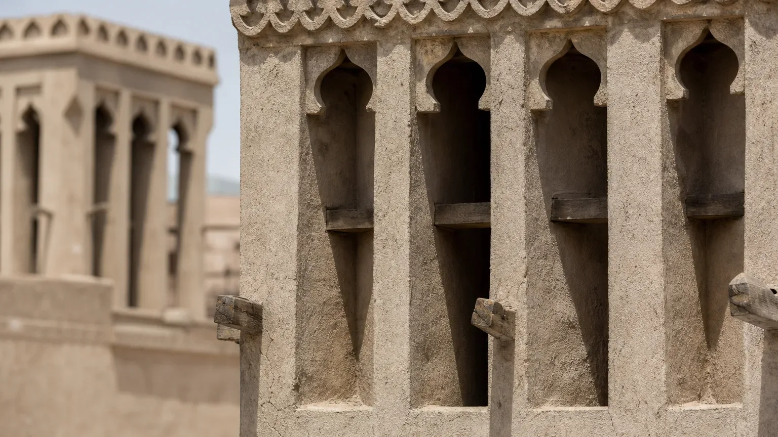 Close-up of ornate mud-plaster wind-tower openings with carved arched vents and wooden beams, a second similar structure blurred in the background