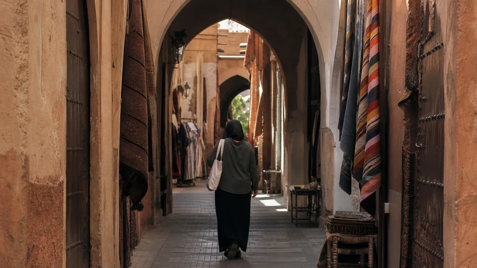 A person carrying a tote bag walks away from the camera along a narrow covered arcade lined with hanging rugs and textiles