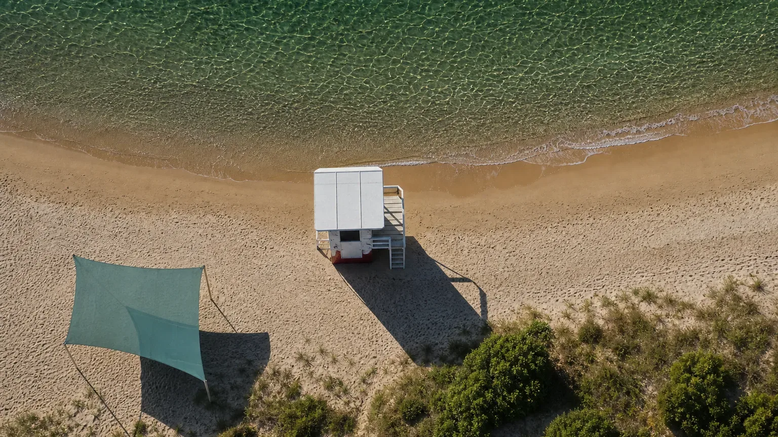 An aerial view of a white lifeguard station and a teal shade canopy on a sandy beach beside clear shallow water, with no people present