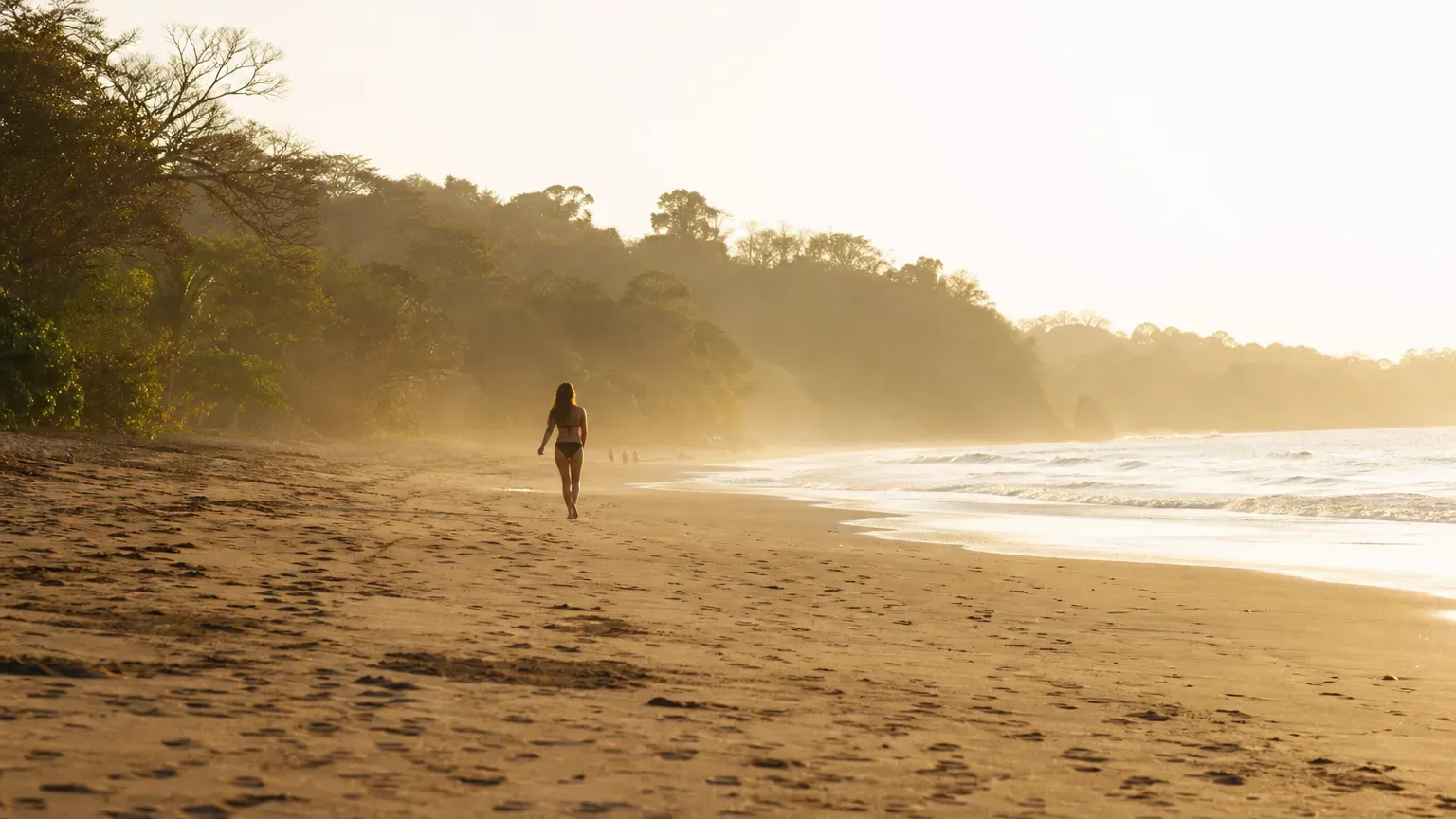 A person in swimwear walks along a wide, empty sandy beach beside gently breaking waves, with dense tropical trees lining the shore in warm golden backlit haze