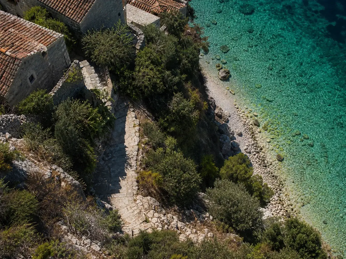 Ancient stone buildings and a stepped cobbled path wind down through dense scrub to a clear turquoise pebble cove, viewed from directly overhead in strong midday sun