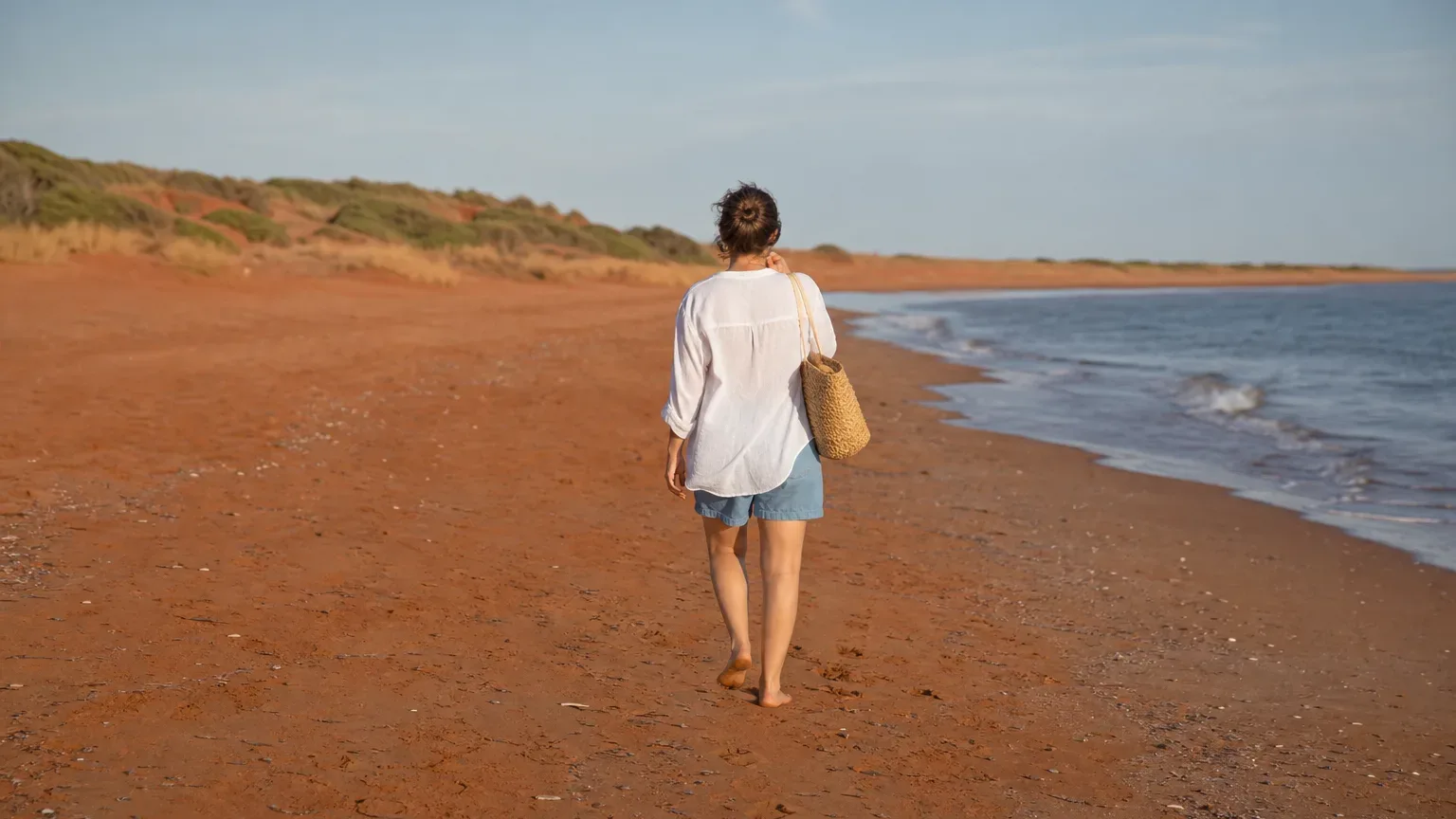 A person walks barefoot along a rust-red sandy beach beside gentle waves, carrying a woven bag, with low dunes and scrub behind them