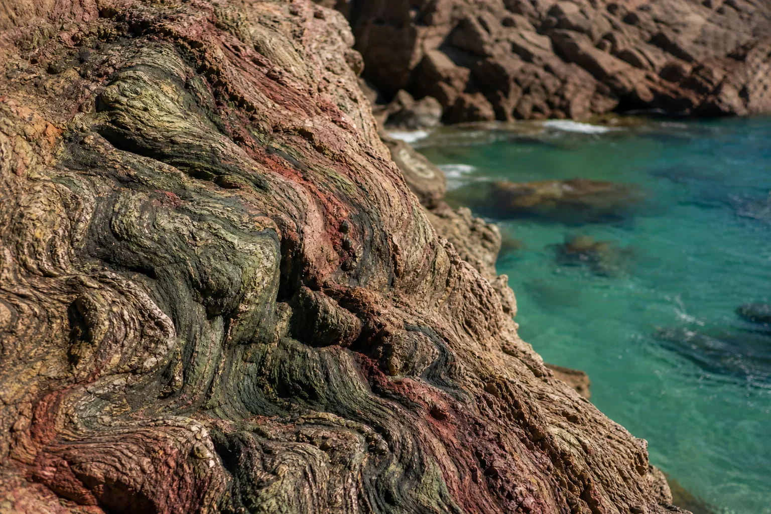 Close-up of dramatically folded serpentine rock in swirling green, red and black beside clear turquoise coastal water