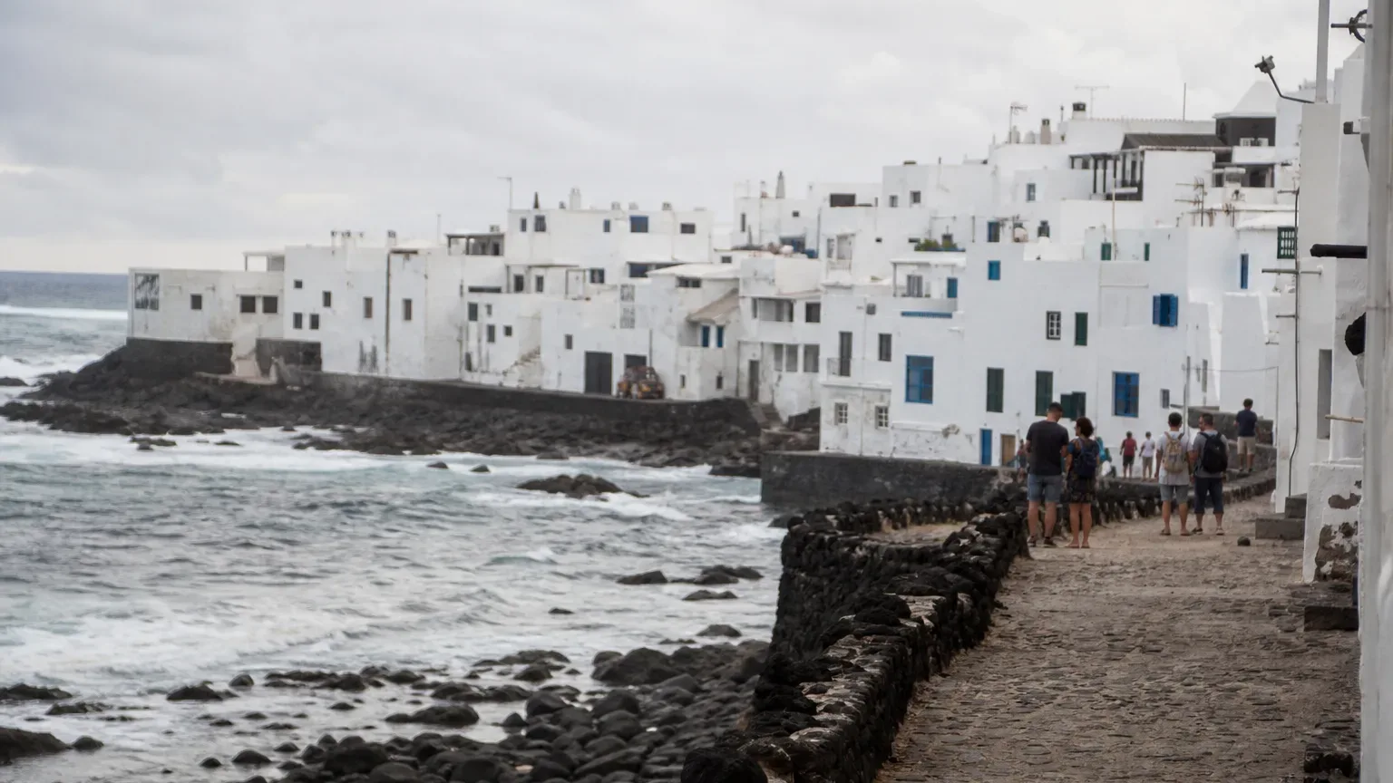 A group of people walk along a cobbled seafront path beside a low stone sea wall, with whitewashed buildings rising behind them and Atlantic waves breaking on dark rocks below