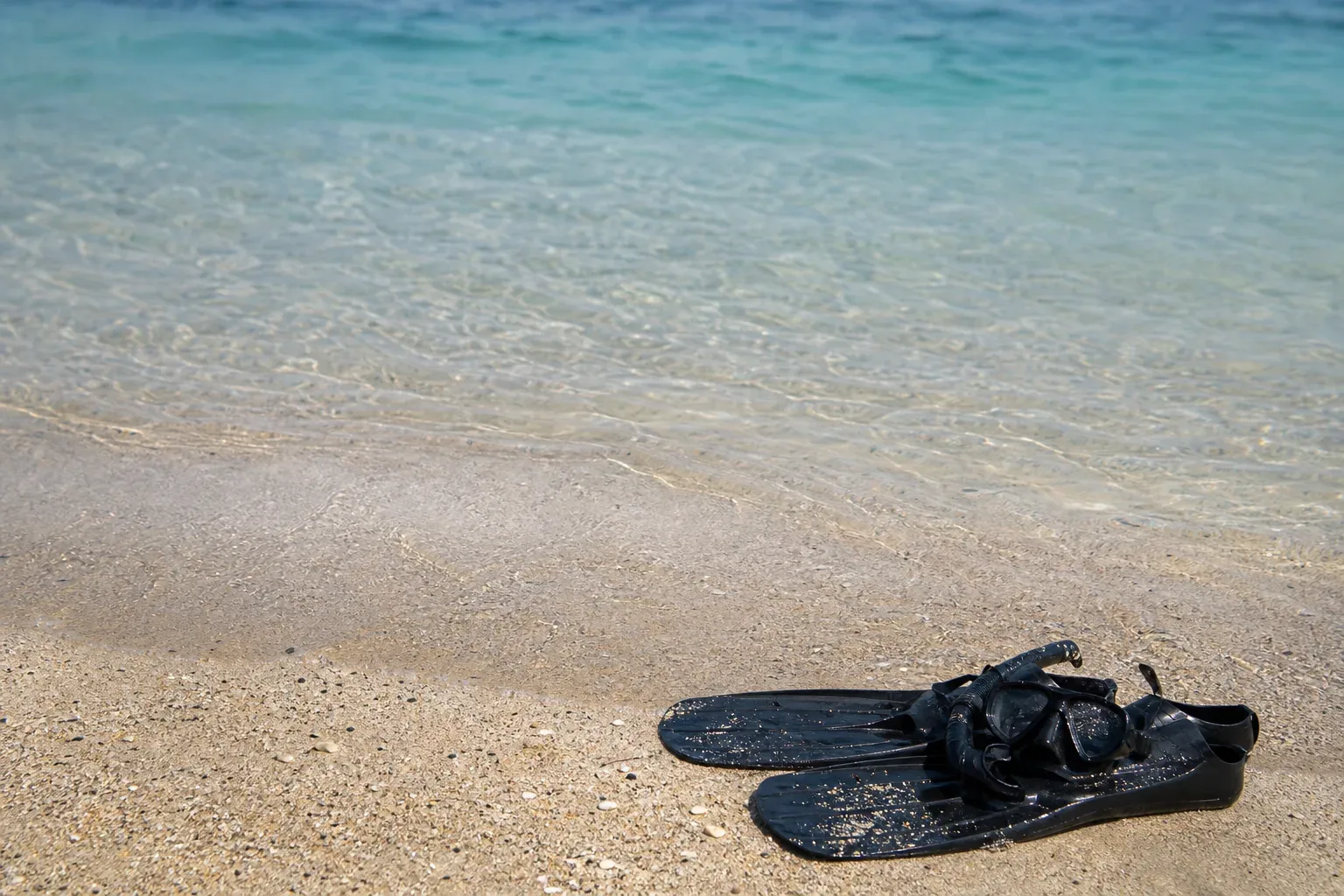 A pair of black swimming fins and a snorkel mask rest on wet sand at the edge of a calm, clear-watered shoreline