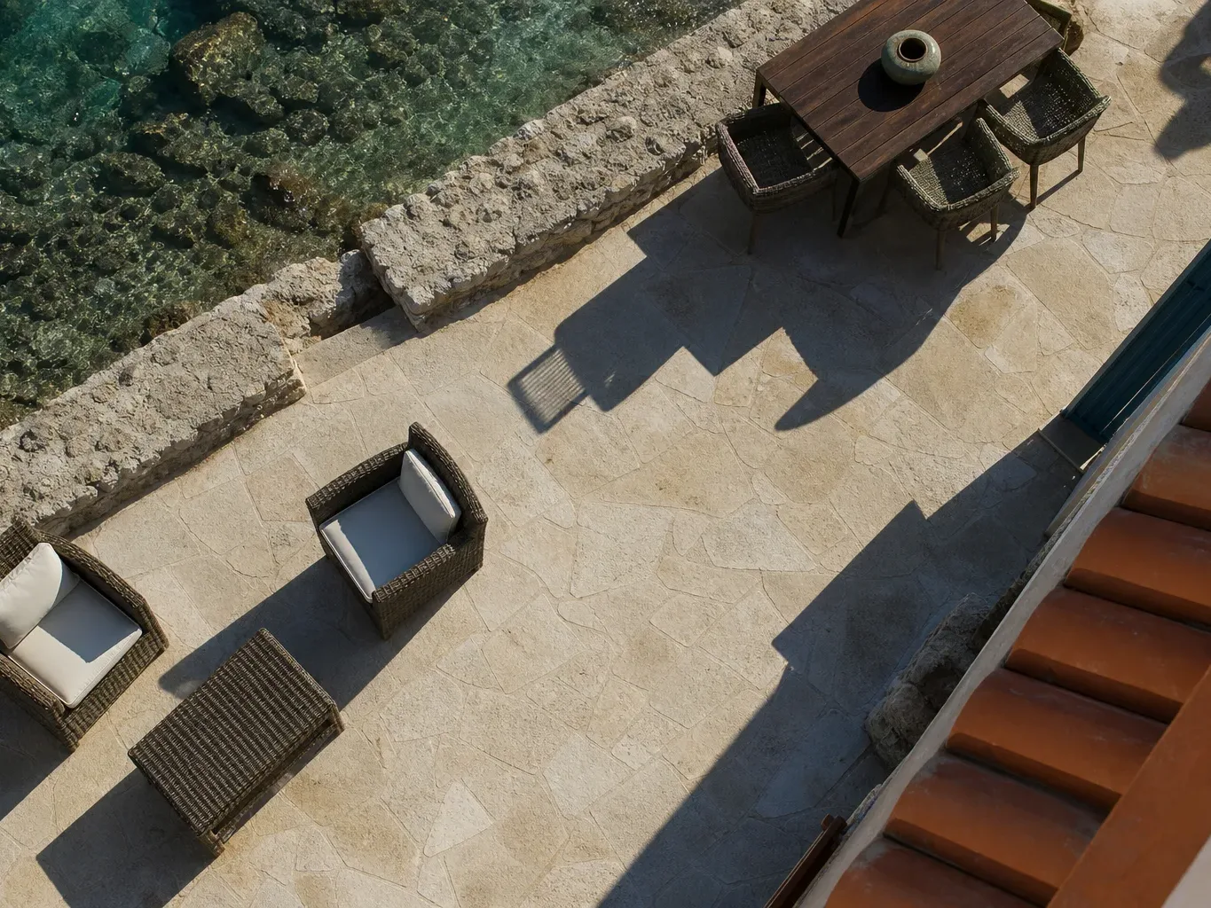 An overhead view of a stone-paved waterside terrace with wicker armchairs, a dining table and chairs, and turquoise water beyond a rocky seawall