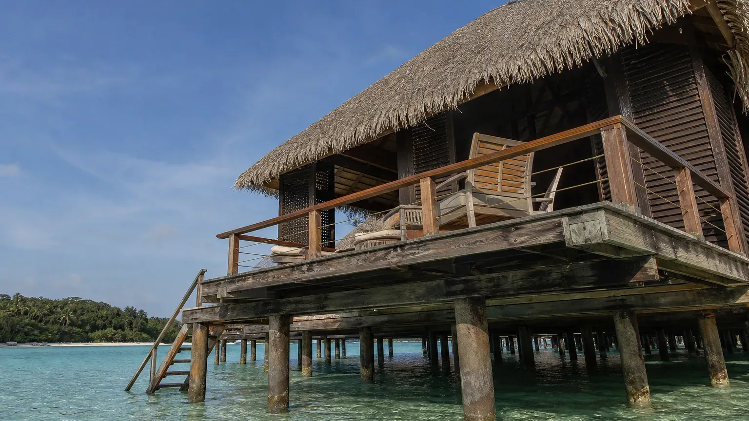 A thatched overwater bungalow on timber stilts above clear turquoise water, with a wooden deck and lounge chairs, a ladder descending to the water, and a palm-fringed shore in the distance