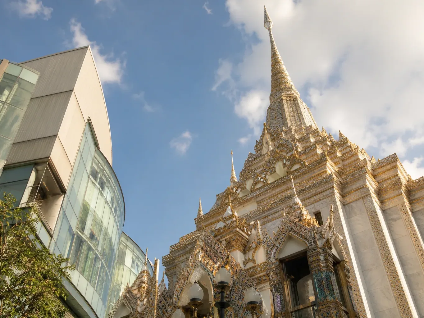 An ornate white-and-gold temple spire rises beside a curved glass-fronted modern building beneath a partly cloudy blue sky, viewed from street level looking upward