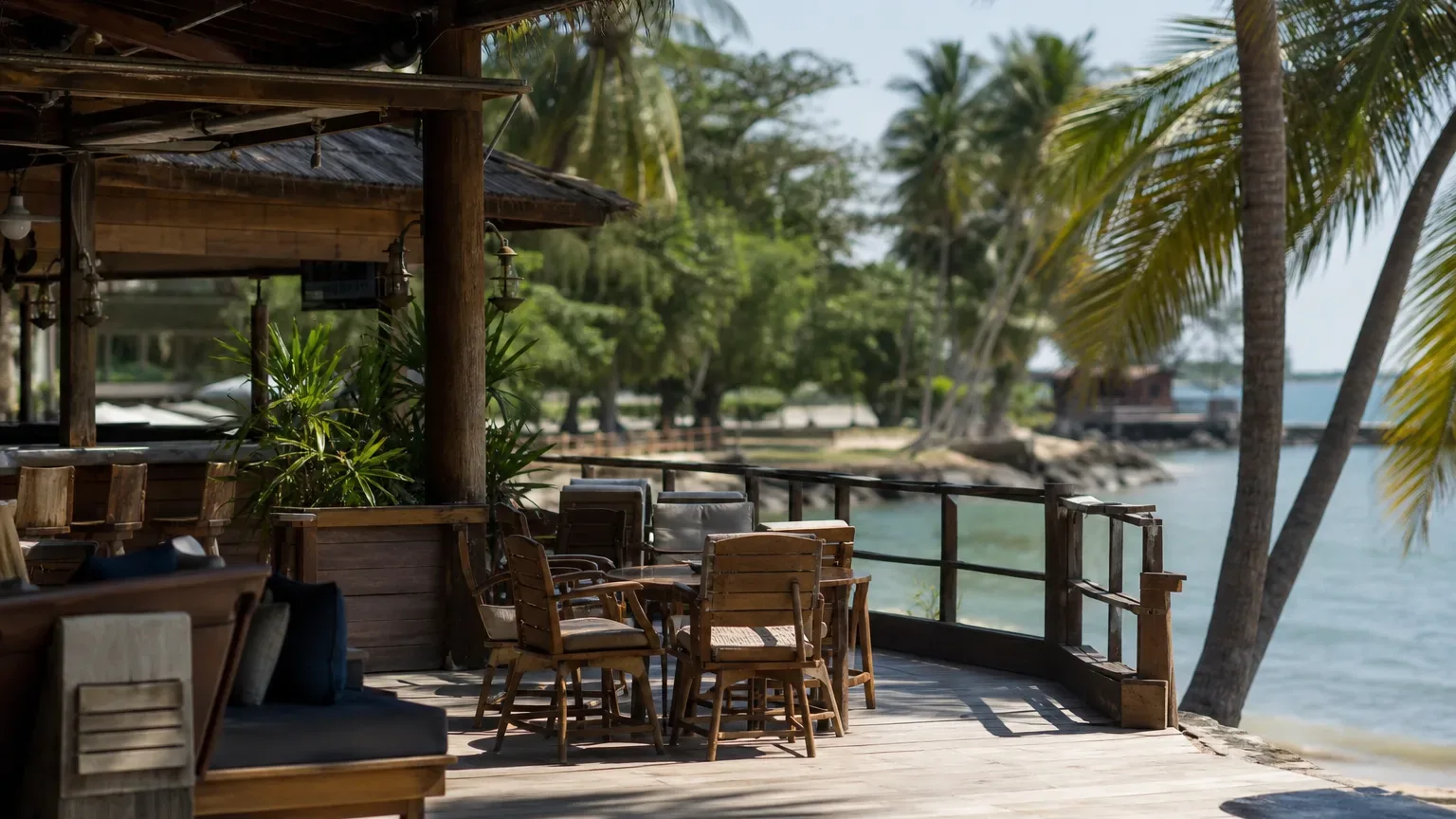 A wooden open-air beach bar and terrace with timber chairs and tables overlooks calm tropical water fringed by palm trees, with no one present