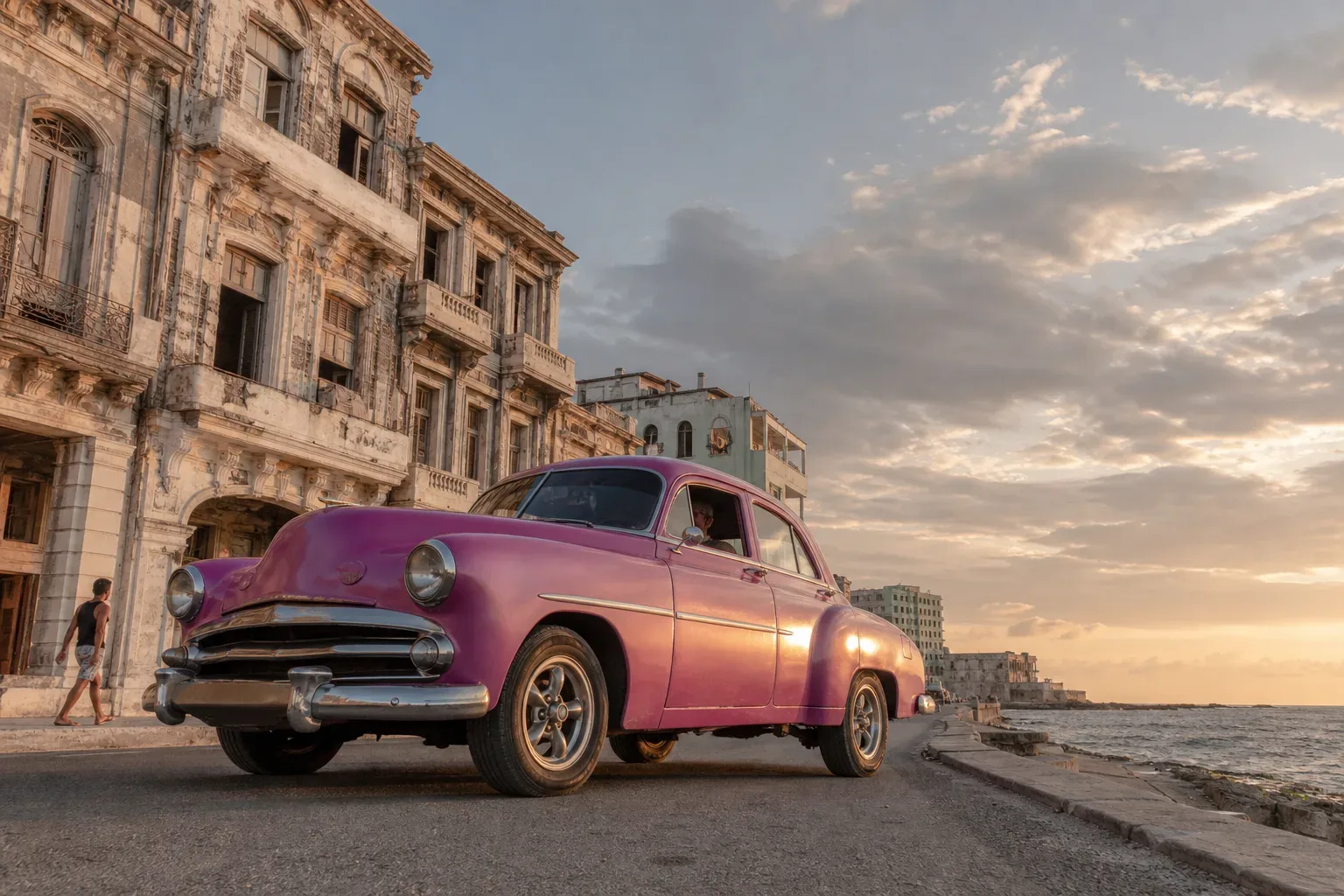A pink vintage saloon car drives along a coastal road beside weathered neoclassical building façades at sunset, with a person walking on the pavement behind