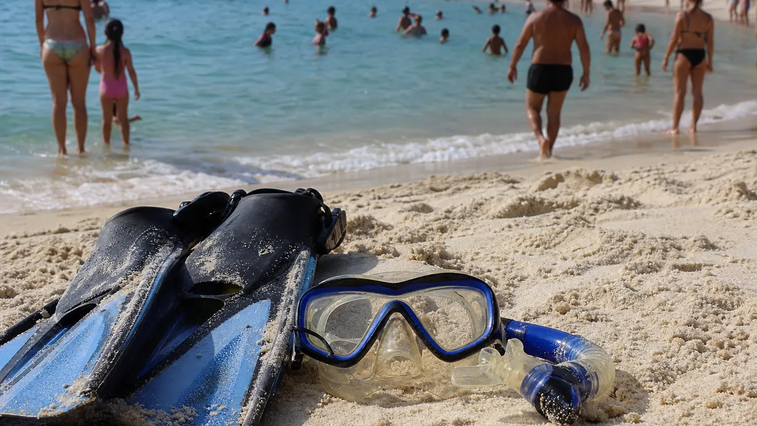 A snorkel mask and fins lying on pale sand in the foreground, with a crowded beach and calm sea filled with swimmers behind