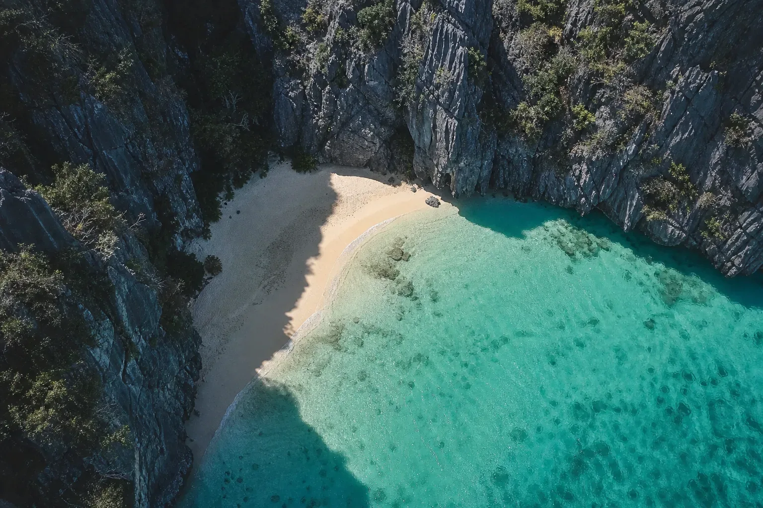 An aerial view of a secluded crescent beach of pale sand enclosed by tall dark limestone cliffs, with vivid turquoise shallow water revealing coral beneath the surface