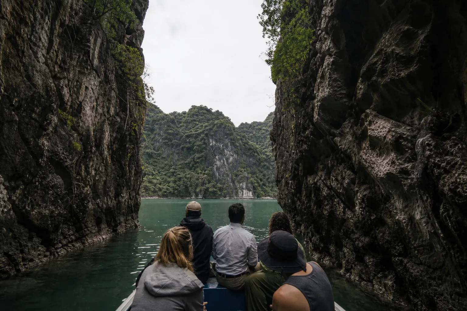 A small group of people sit at the front of a boat passing through a narrow gap between tall karst rock formations, with a lush green island visible ahead on calm water