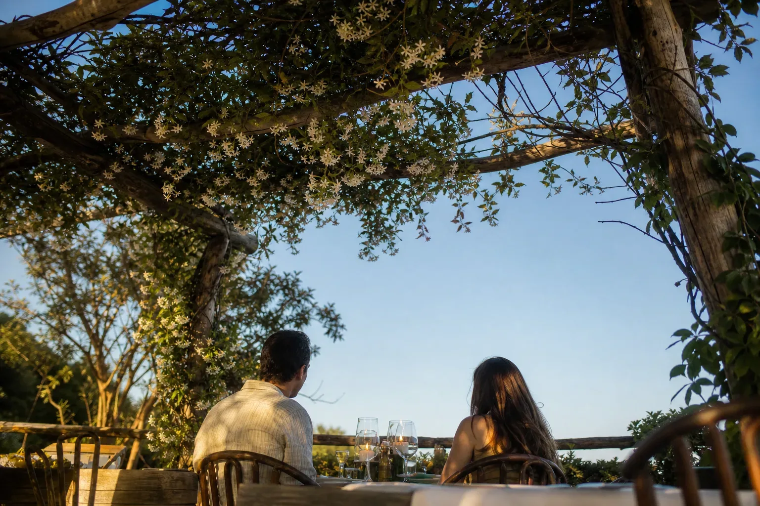 Two people sit with their backs to the camera at a candlelit outdoor dining table beneath a rustic pergola draped in white flowering vines, with open evening sky beyond