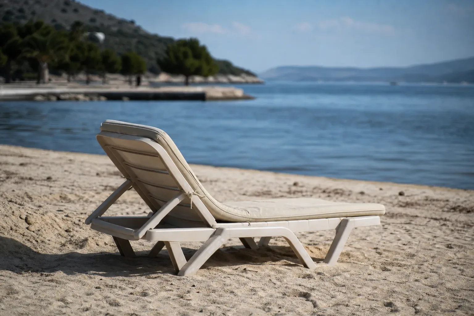 An empty sun-lounger with a cushioned pad sits on pale sandy shore beside calm blue water, with a tree-lined headland visible in the background