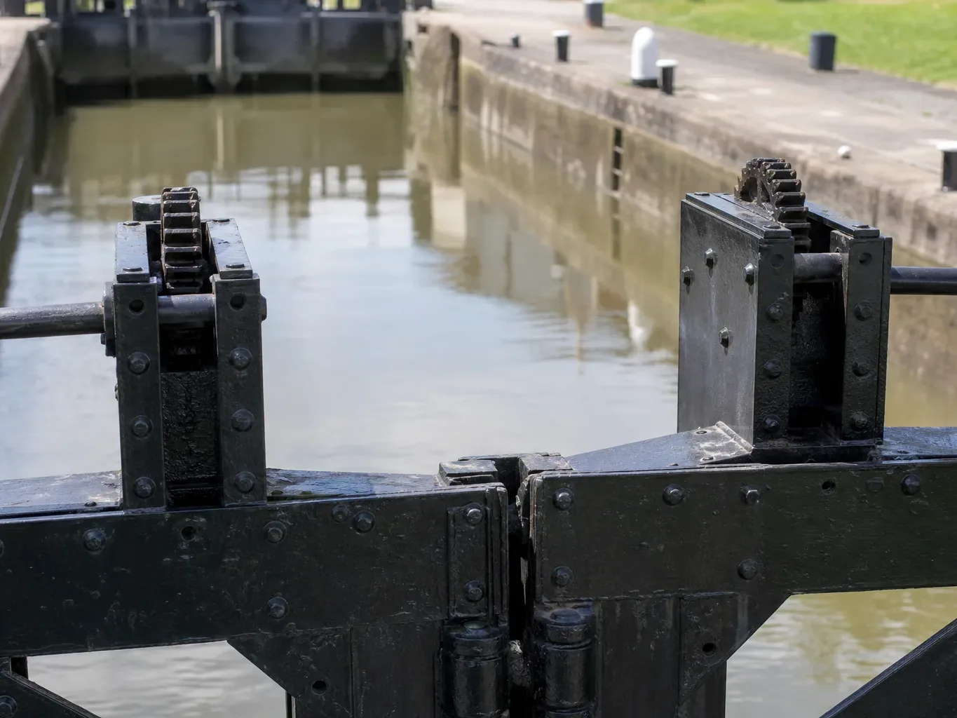 Close-up of cast-iron rack-and-pinion gate mechanisms on a canal lock, with the empty lock chamber and concrete towpath visible behind