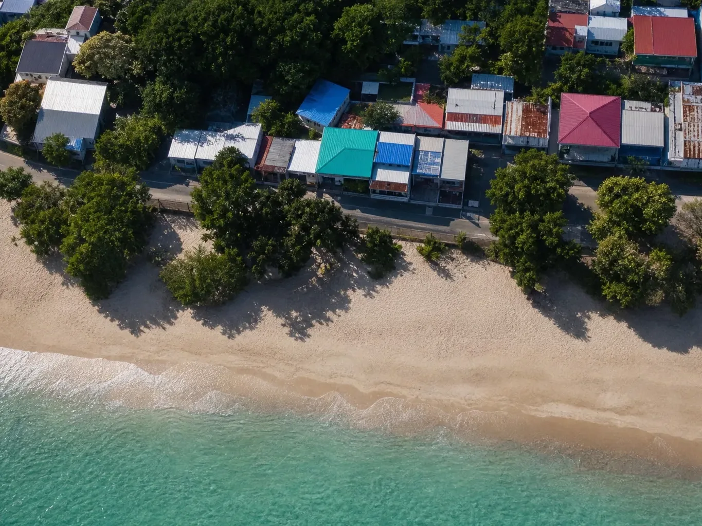 An aerial view of a sandy beach with calm turquoise water lapping the shore, backed by a tree-lined coastal road and a cluster of colourful metal-roofed buildings