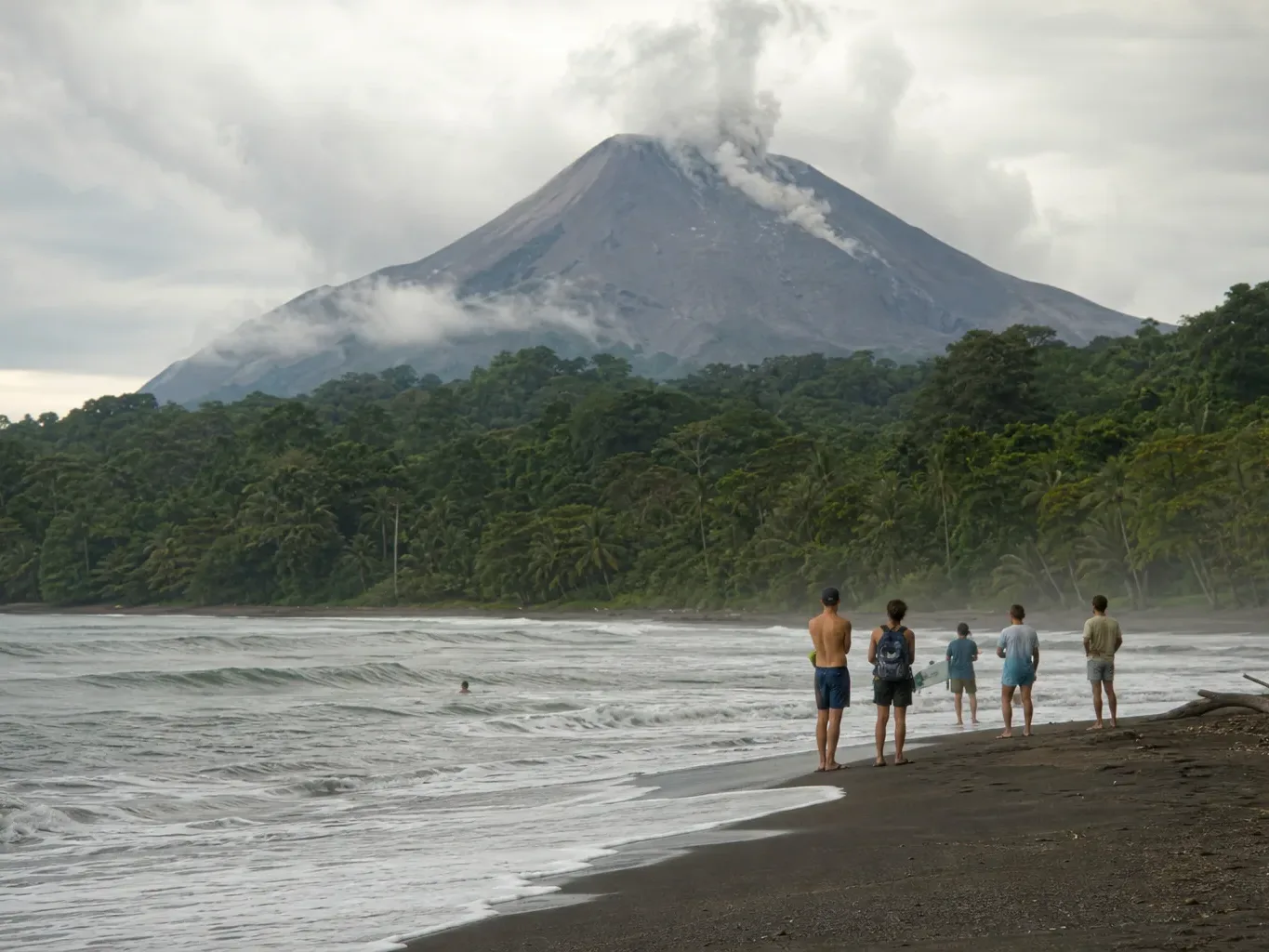 A small group of people stand at the shoreline of a dark-sand beach watching waves, with dense rainforest and an active volcano emitting smoke and steam rising behind