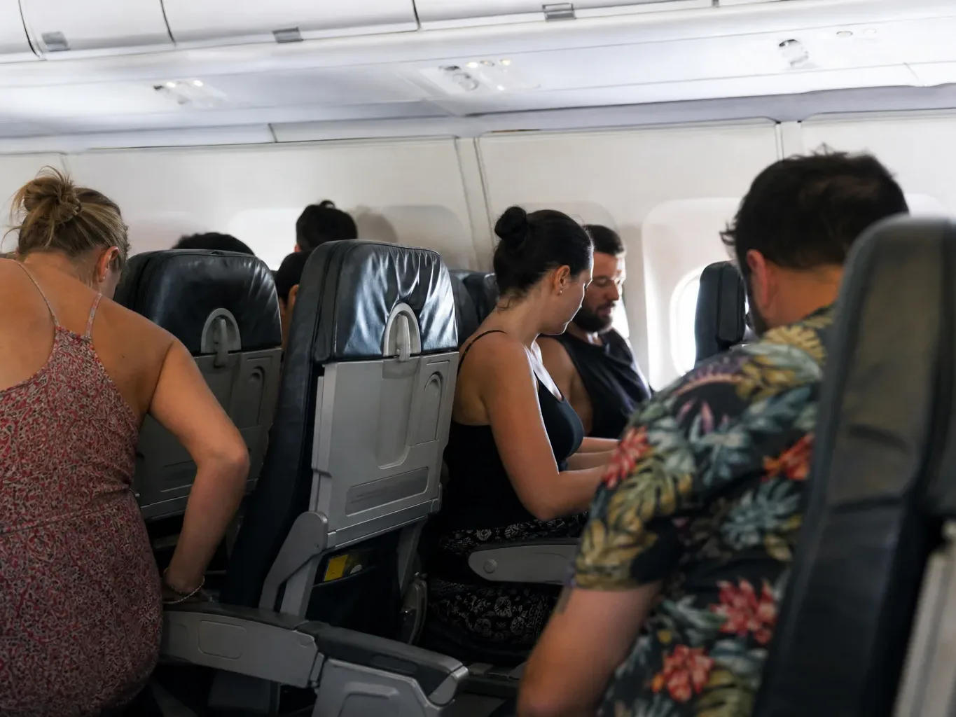 Passengers seated in rows of economy seats inside an aircraft cabin, some looking down as they settle in