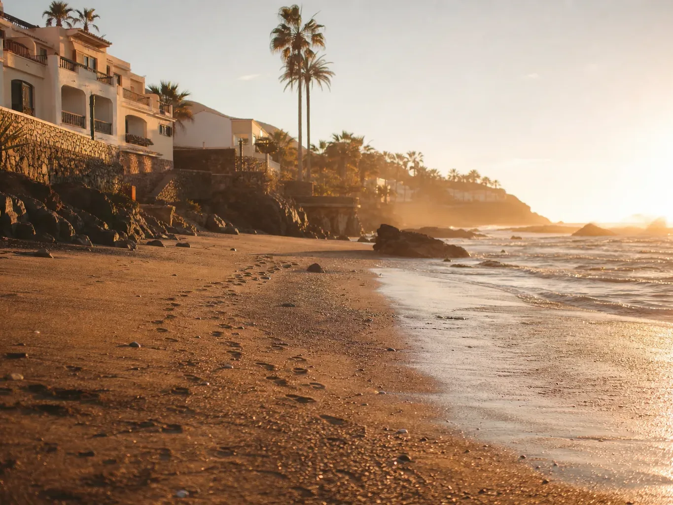 A sandy beach at golden hour, with gentle waves lapping the shore, rocky outcrops, whitewashed buildings on a terraced hillside, and tall palm trees silhouetted against a warm hazy sky